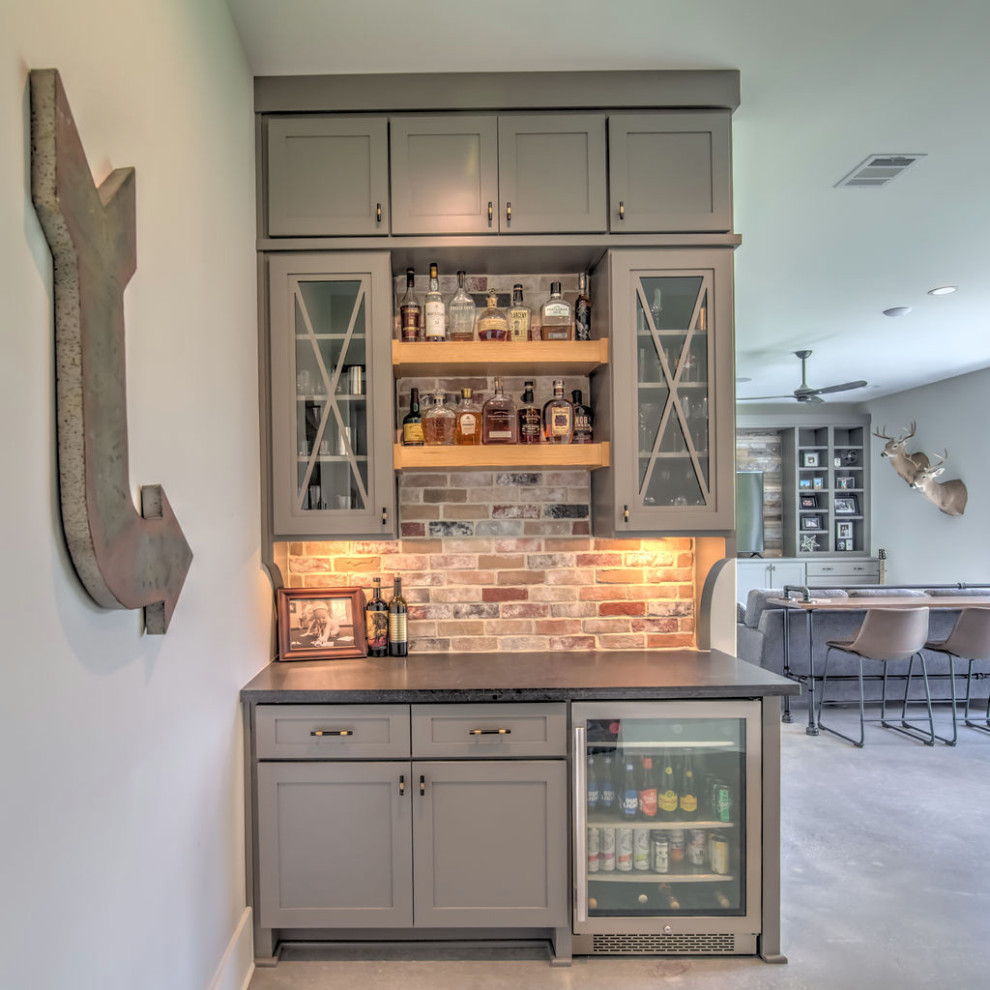 This is an example of a small country single-wall home bar in Houston with recessed-panel cabinets, grey cabinets, multi-coloured splashback, brick splashback, concrete floors, grey floor and brown benchtop.