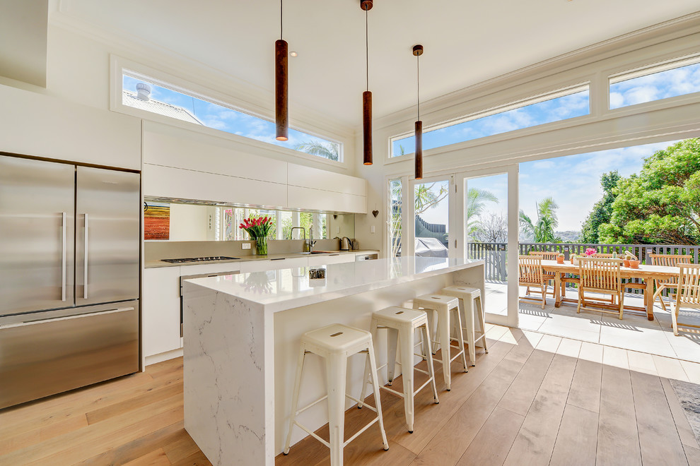 Inspiration for a large contemporary galley open plan kitchen in Sydney with a drop-in sink, white cabinets, quartz benchtops, mirror splashback, stainless steel appliances, with island, flat-panel cabinets, brown floor, metallic splashback, medium hardwood floors and white benchtop.