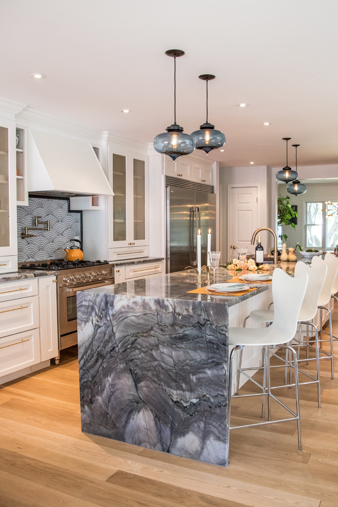 Large transitional open plan kitchen with an undermount sink, white cabinets, quartzite benchtops, glass tile splashback, stainless steel appliances, light hardwood floors, with island, shaker cabinets, blue splashback and brown floor.