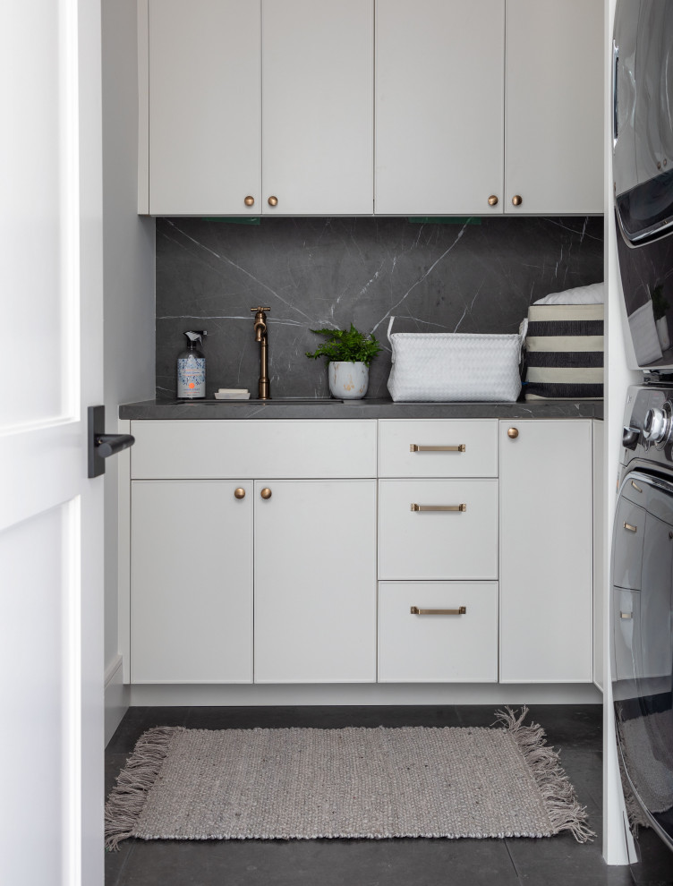 This is an example of a mid-sized transitional u-shaped dedicated laundry room in Houston with a drop-in sink, flat-panel cabinets, grey cabinets, white walls, a stacked washer and dryer and grey benchtop.