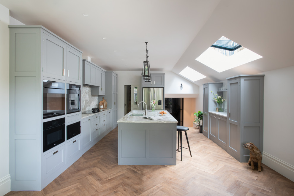 Mid-sized transitional u-shaped eat-in kitchen in London with an undermount sink, shaker cabinets, grey cabinets, marble benchtops, multi-coloured splashback, marble splashback, black appliances, medium hardwood floors, with island and multi-coloured benchtop.