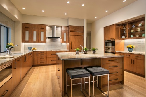 Modern kitchen cabinet design in warm wood with flat-panel doors, large island and stools, built-in ovens, and open display shelves.