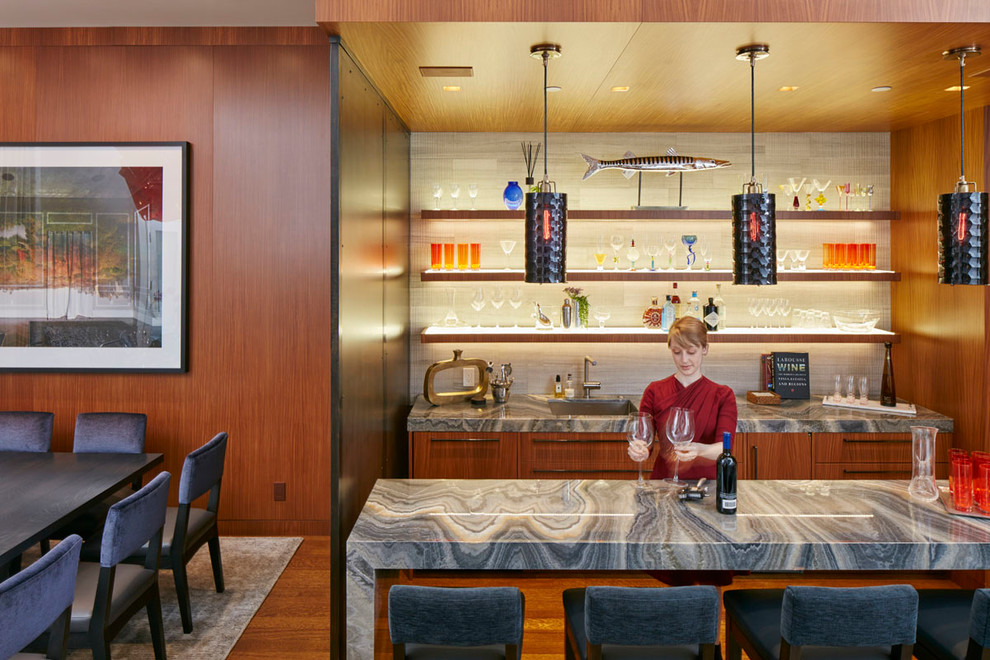 This is an example of a mid-sized contemporary galley seated home bar in New York with an undermount sink, flat-panel cabinets, medium wood cabinets, onyx benchtops, beige splashback, stone tile splashback, medium hardwood floors, brown floor and grey benchtop.