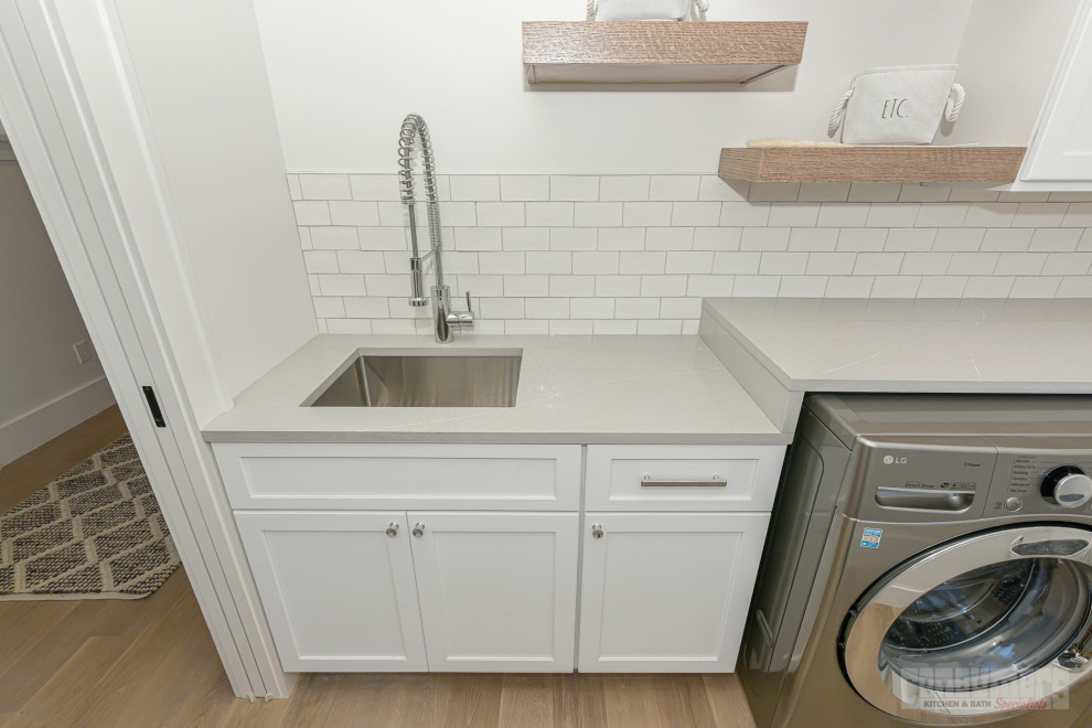 Retro inredning av en liten grå linjär grått tvättstuga enbart för tvätt, med en nedsänkt diskho, luckor med infälld panel, vita skåp, bänkskiva i kvarts, vitt stänkskydd, stänkskydd i tunnelbanekakel, vita väggar, mellanmörkt trägolv, en tvättmaskin och torktumlare bredvid varandra och brunt golv