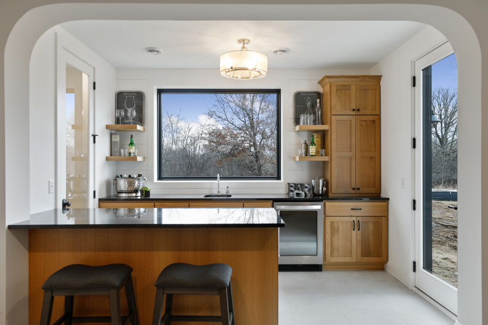 Design ideas for a transitional galley seated home bar in Minneapolis with an undermount sink, shaker cabinets, medium wood cabinets, white splashback, white floor and black benchtop.