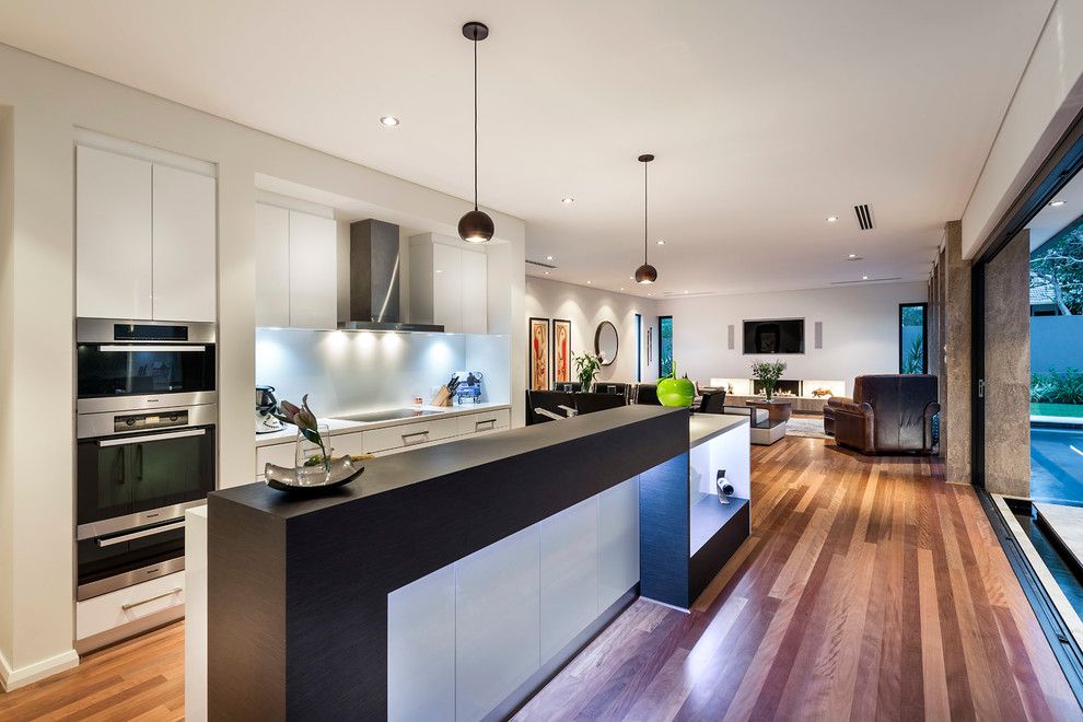 Design ideas for a mid-sized contemporary galley open plan kitchen in Perth with flat-panel cabinets, white cabinets, white splashback, glass sheet splashback, stainless steel appliances, medium hardwood floors and with island.