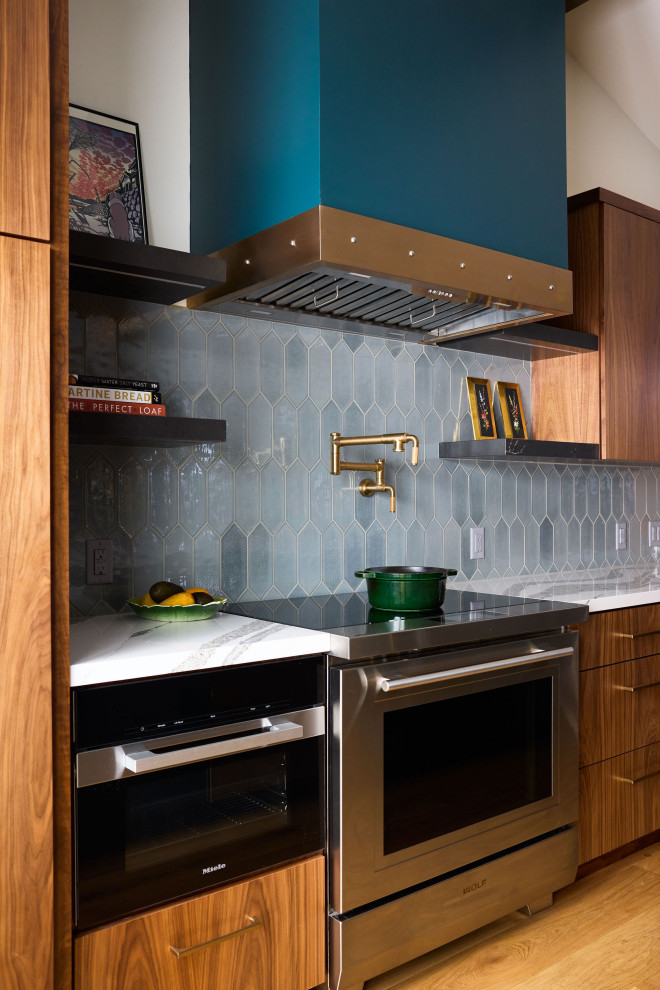 Example of a large eclectic medium tone wood floor, brown floor and vaulted ceiling kitchen design in Other with a drop-in sink, flat-panel cabinets, medium tone wood cabinets, quartz countertops, blue backsplash, ceramic backsplash, stainless steel appliances, an island and white countertops
