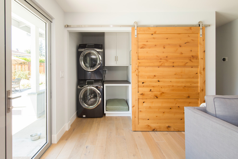 Small transitional single-wall laundry cupboard in San Francisco with white cabinets, light hardwood floors, a stacked washer and dryer, flat-panel cabinets, solid surface benchtops and grey benchtop.