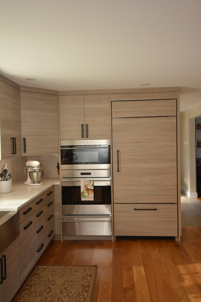 This is an example of a mid-sized contemporary l-shaped kitchen pantry in Burlington with a farmhouse sink, flat-panel cabinets, light wood cabinets, quartz benchtops, grey splashback, ceramic splashback, stainless steel appliances, medium hardwood floors, a peninsula, brown floor and white benchtop.