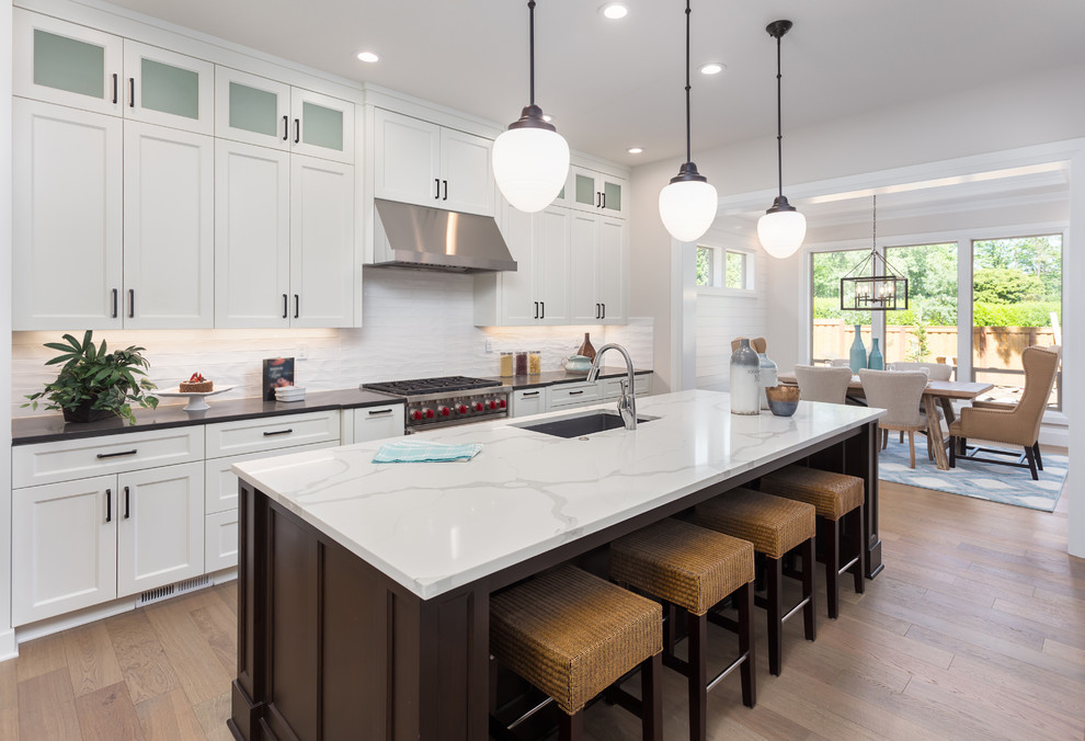 Design ideas for a transitional l-shaped eat-in kitchen in DC Metro with an undermount sink, shaker cabinets, white cabinets, white splashback, subway tile splashback, medium hardwood floors, with island, brown floor and white benchtop.