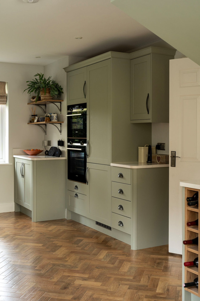 Design ideas for a mid-sized transitional single-wall kitchen pantry in Oxfordshire with a single-bowl sink, raised-panel cabinets, green cabinets, marble benchtops, white splashback, cement tile splashback, black appliances, medium hardwood floors, no island, brown floor and white benchtop.