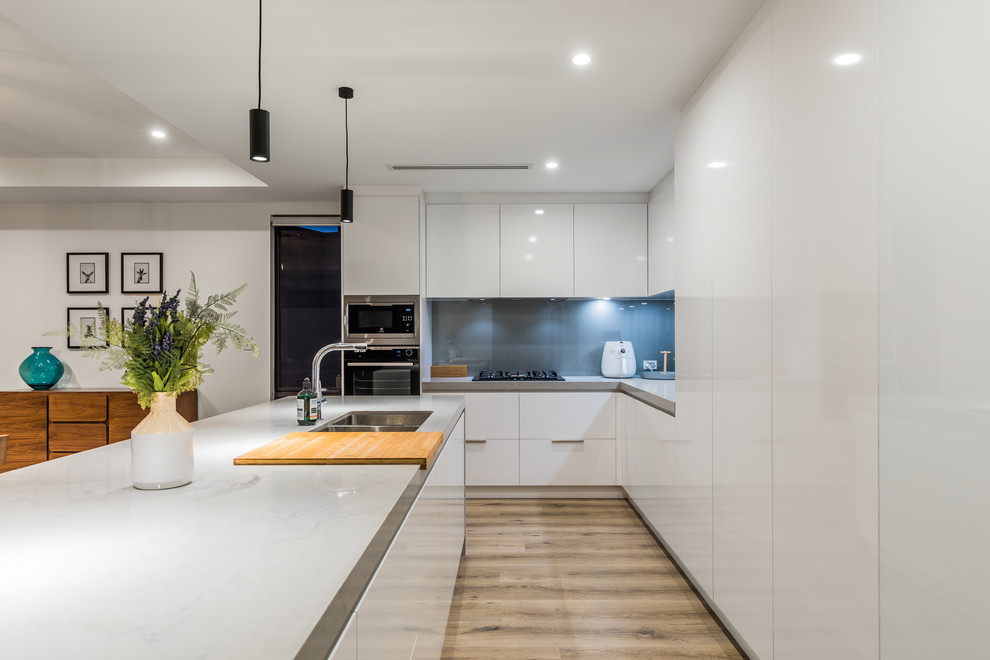 Photo of a contemporary l-shaped kitchen in Perth with an undermount sink, flat-panel cabinets, white cabinets, blue splashback, glass sheet splashback, medium hardwood floors, with island, brown floor and grey benchtop.