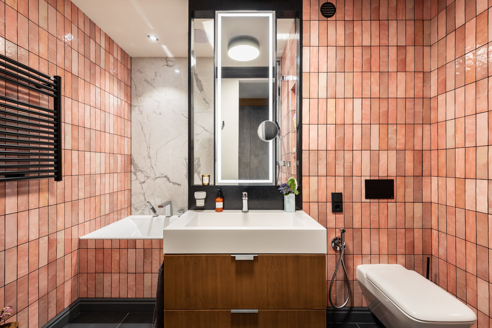 Inspiration for a contemporary bathroom in Saint Petersburg with flat-panel cabinets, medium wood cabinets, an undermount tub, a wall-mount toilet, pink tile, ceramic tile, pink walls, porcelain floors, black floor and an integrated sink.