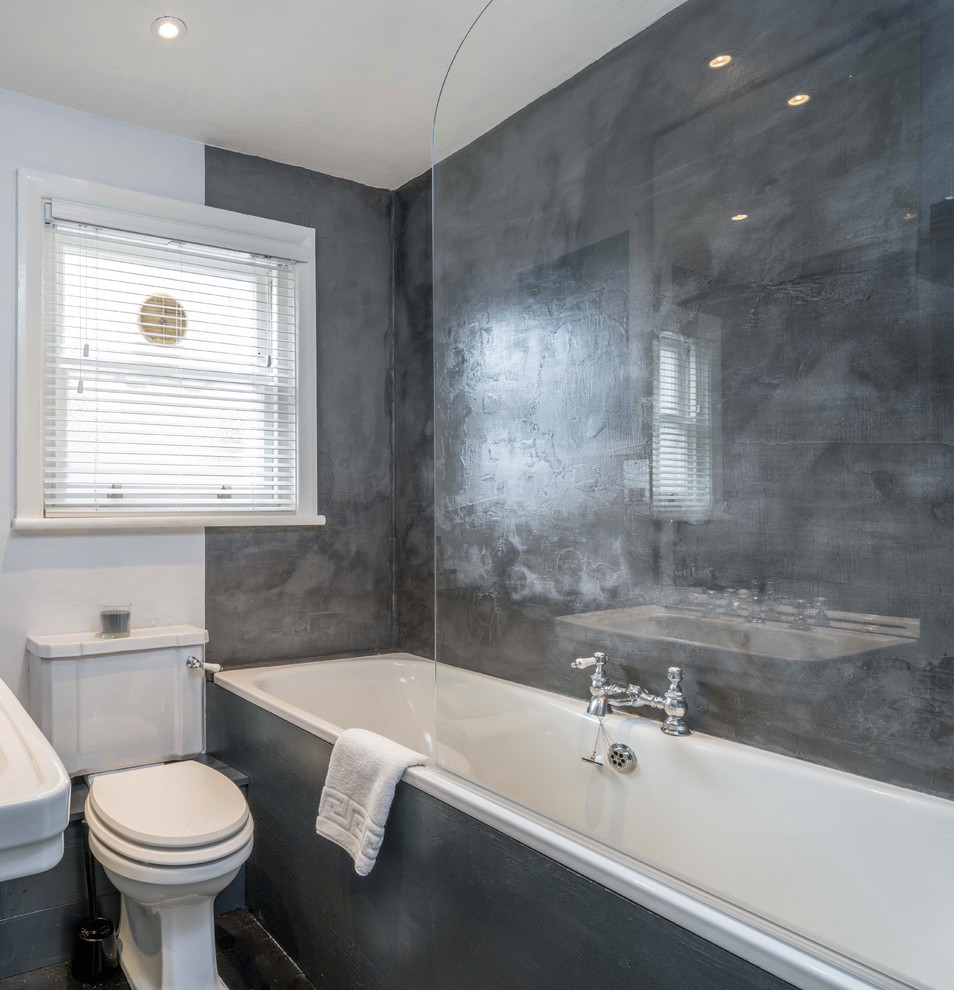 This is an example of a mid-sized transitional kids bathroom in London with a drop-in tub, a shower/bathtub combo, a one-piece toilet, gray tile, cement tile, grey walls, painted wood floors and concrete benchtops.