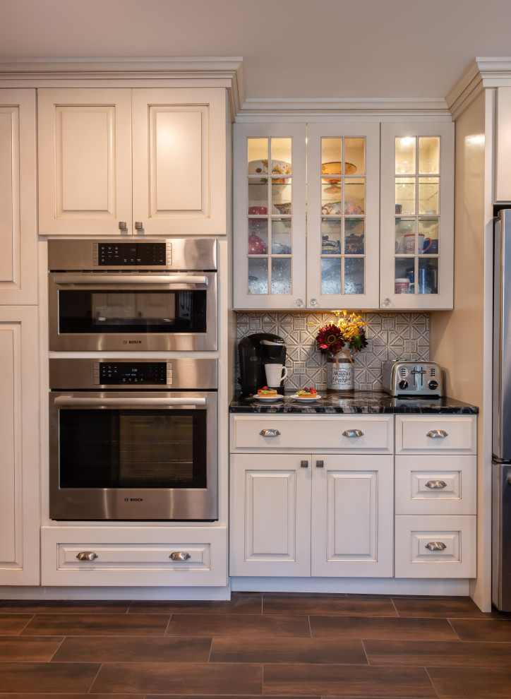 This is an example of a large transitional l-shaped eat-in kitchen in Other with a farmhouse sink, raised-panel cabinets, distressed cabinets, granite benchtops, beige splashback, glass tile splashback, stainless steel appliances, porcelain floors, with island, brown floor and black benchtop.