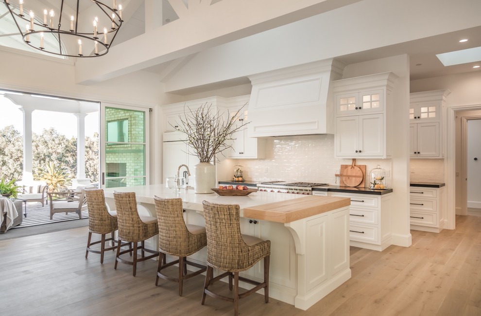 This is an example of a country kitchen in San Diego with shaker cabinets, white cabinets, white splashback, stainless steel appliances, light hardwood floors, with island, beige floor and black benchtop.