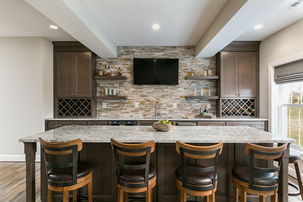 Photo of a large transitional galley seated home bar in DC Metro with recessed-panel cabinets, brown cabinets, granite benchtops, beige splashback, stone tile splashback, laminate floors, brown floor, an undermount sink and grey benchtop.