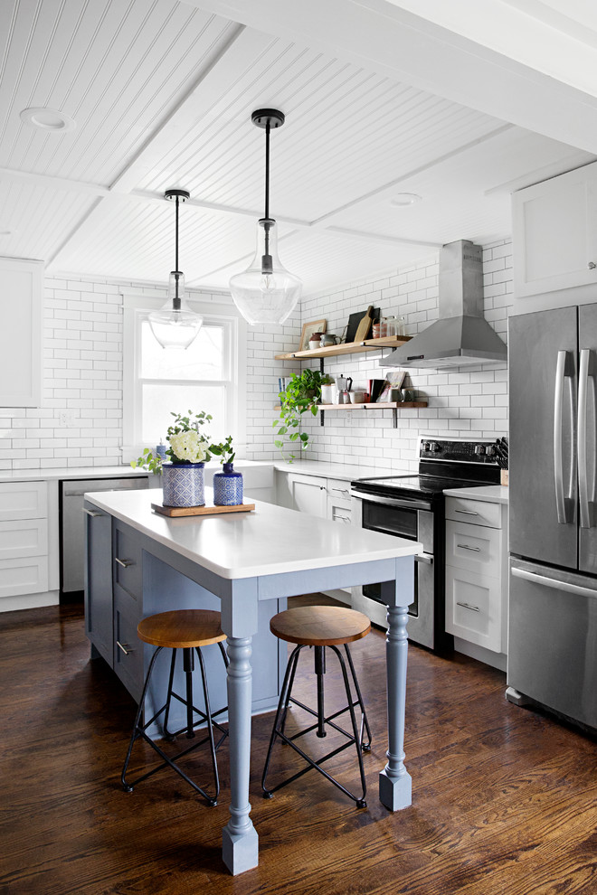 This is an example of a transitional l-shaped kitchen in Nashville with a farmhouse sink, shaker cabinets, white cabinets, white splashback, subway tile splashback, stainless steel appliances, medium hardwood floors, with island, brown floor and white benchtop.