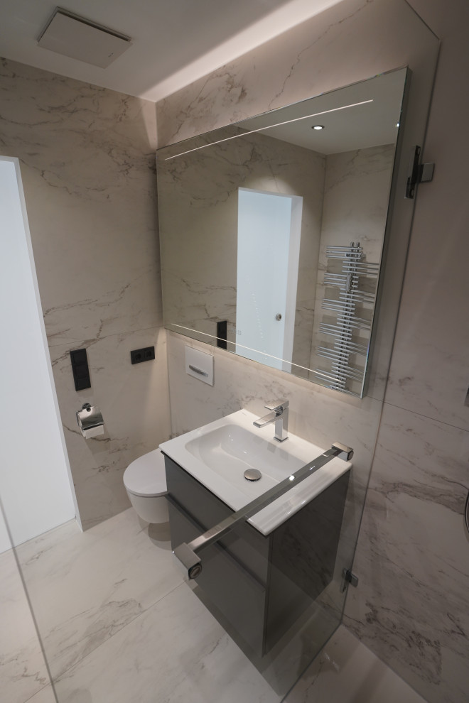 Bild på ett mellanstort funkis vit vitt badrum med dusch, med släta luckor, grå skåp, en hörndusch, en vägghängd toalettstol, svart och vit kakel, stenkakel, vita väggar, ett väggmonterat handfat, vitt golv och med dusch som är öppen