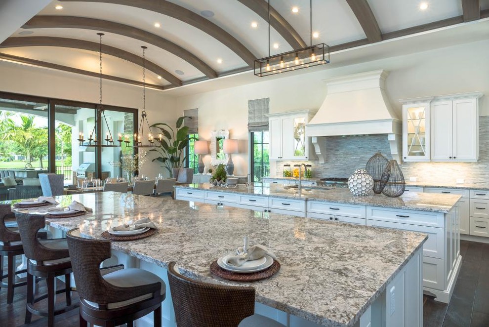 Large mediterranean u-shaped eat-in kitchen in Miami with a farmhouse sink, recessed-panel cabinets, white cabinets, granite benchtops, grey splashback, stone tile splashback, panelled appliances, dark hardwood floors, multiple islands and brown floor.