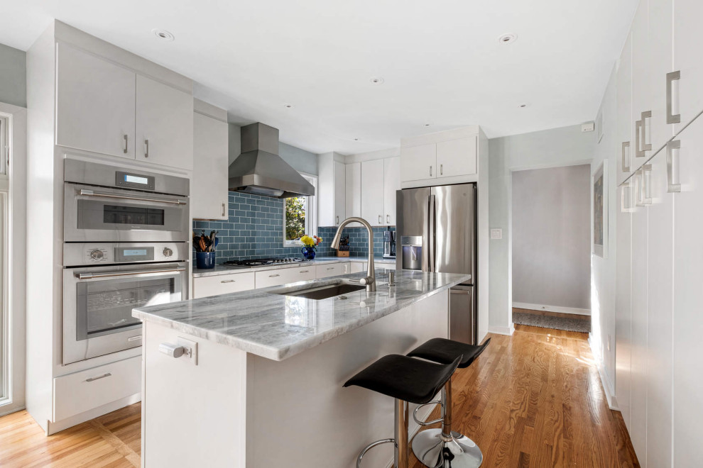 Photo of a mid-sized contemporary l-shaped eat-in kitchen in Philadelphia with flat-panel cabinets, white cabinets, blue splashback, glass tile splashback, stainless steel appliances, medium hardwood floors, with island, an undermount sink, granite benchtops, brown floor and grey benchtop.