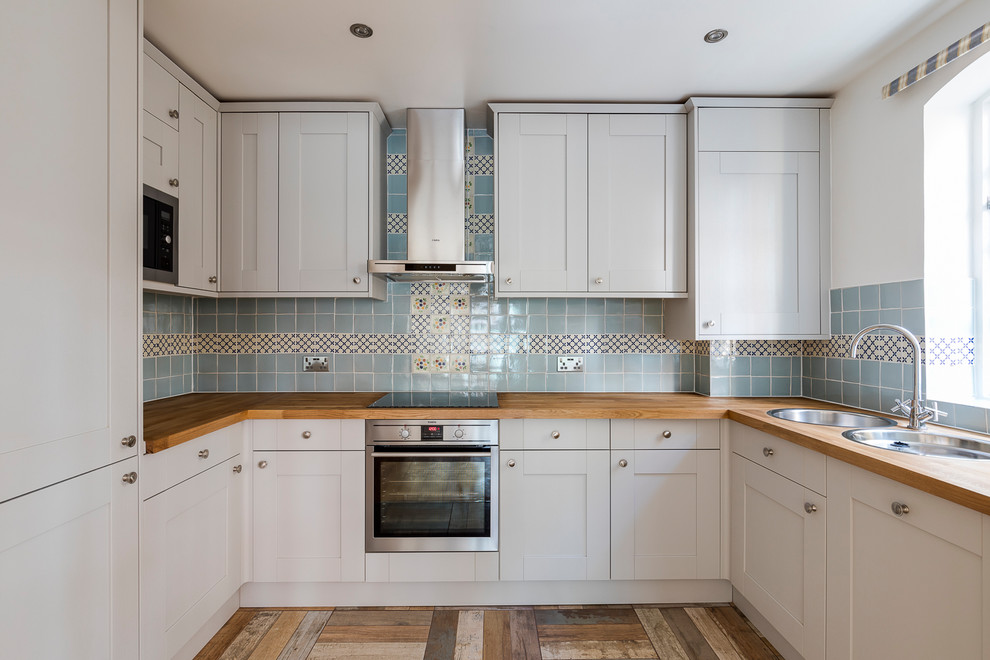 Mid-sized traditional u-shaped eat-in kitchen in London with a double-bowl sink, raised-panel cabinets, grey cabinets, wood benchtops, blue splashback, mosaic tile splashback, black appliances, cement tiles and no island.