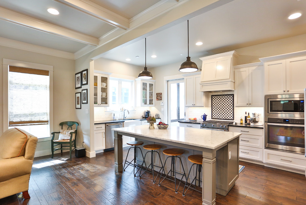 Mid-sized traditional u-shaped open plan kitchen in Austin with a farmhouse sink, recessed-panel cabinets, white cabinets, granite benchtops, black splashback, ceramic splashback, stainless steel appliances, medium hardwood floors, with island, brown floor and white benchtop.
