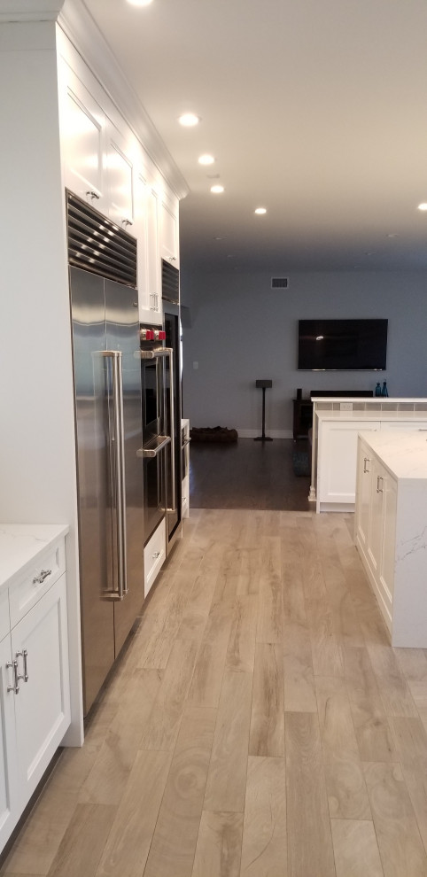 This is an example of a transitional u-shaped eat-in kitchen in New York with a farmhouse sink, recessed-panel cabinets, white cabinets, quartz benchtops, grey splashback, glass tile splashback, stainless steel appliances, porcelain floors, with island, brown floor and multi-coloured benchtop.