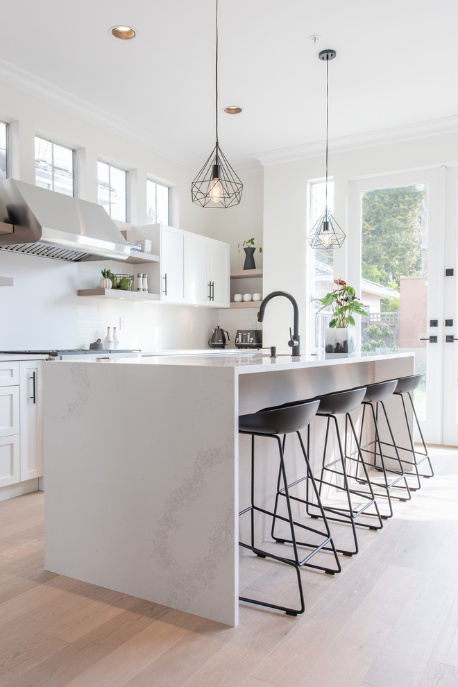 Scandinavian l-shaped kitchen in Vancouver with an undermount sink, shaker cabinets, white cabinets, quartz benchtops, white splashback, stainless steel appliances, light hardwood floors, with island and white benchtop.