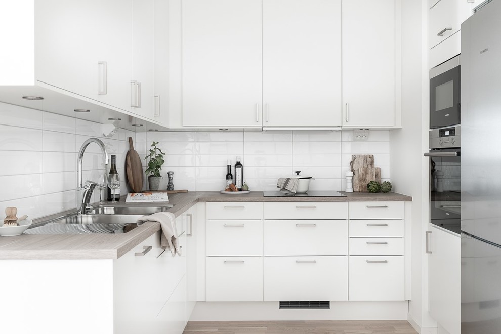 Inspiration for a scandinavian l-shaped kitchen in Stockholm with a double-bowl sink, flat-panel cabinets, white cabinets, wood benchtops, white splashback, stainless steel appliances, light hardwood floors, beige floor and beige benchtop.