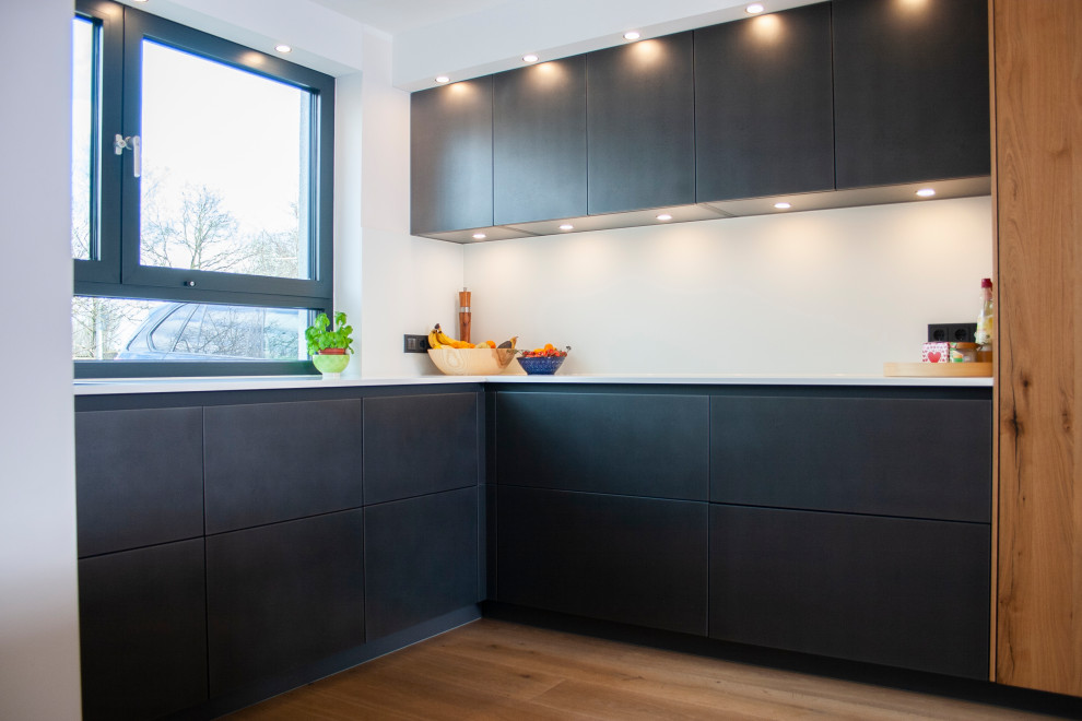 This is an example of a large contemporary l-shaped kitchen/diner in Munich with a submerged sink, flat-panel cabinets, grey cabinets, white splashback, glass sheet splashback, black appliances, painted wood flooring, an island and white worktops.