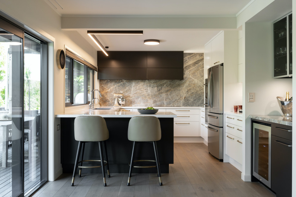This is an example of a mid-sized contemporary u-shaped kitchen in Auckland with a double-bowl sink, flat-panel cabinets, white cabinets, solid surface benchtops, grey splashback, granite splashback, stainless steel appliances, white benchtop, medium hardwood floors, a peninsula and beige floor.