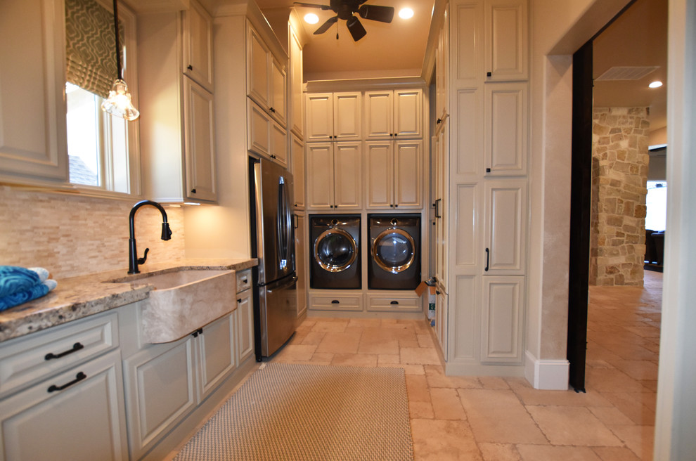 Photo of an expansive mediterranean u-shaped laundry room in Houston with a farmhouse sink, raised-panel cabinets, beige cabinets, granite benchtops, beige walls, travertine floors and a side-by-side washer and dryer.