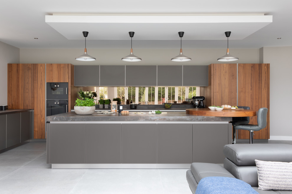 Large contemporary galley open plan kitchen in Hampshire with grey cabinets, quartzite benchtops, metallic splashback, mirror splashback, black appliances, with island, grey benchtop, flat-panel cabinets and grey floor.
