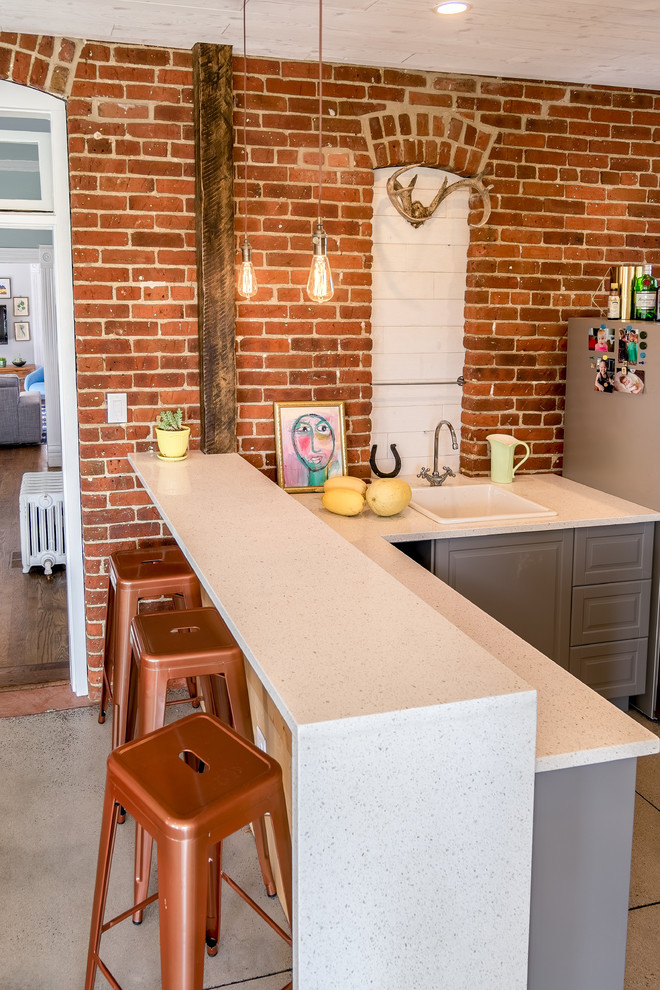 Inspiration for a mid-sized eclectic l-shaped eat-in kitchen in Denver with a drop-in sink, raised-panel cabinets, grey cabinets, quartz benchtops, red splashback, brick splashback, stainless steel appliances, concrete floors, a peninsula and grey floor.