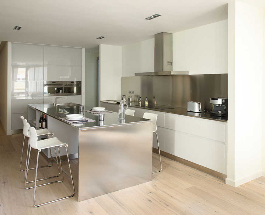 This is an example of a large contemporary single-wall open plan kitchen in Barcelona with an integrated sink, flat-panel cabinets, white cabinets, stainless steel benchtops, grey splashback, stainless steel appliances, light hardwood floors, with island, brown floor and grey benchtop.