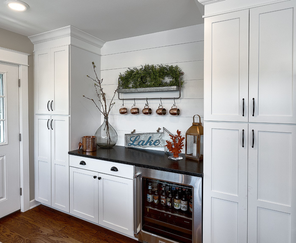 Design ideas for a mid-sized country single-wall open plan kitchen in Chicago with a farmhouse sink, shaker cabinets, white cabinets, granite benchtops, white splashback, ceramic splashback, stainless steel appliances, medium hardwood floors, brown floor and black benchtop.