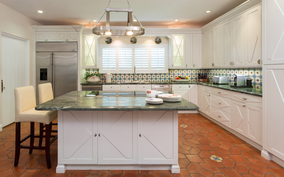 Photo of a mediterranean l-shaped kitchen in Miami with a drop-in sink, recessed-panel cabinets, white cabinets, multi-coloured splashback, stainless steel appliances, terra-cotta floors and with island.
