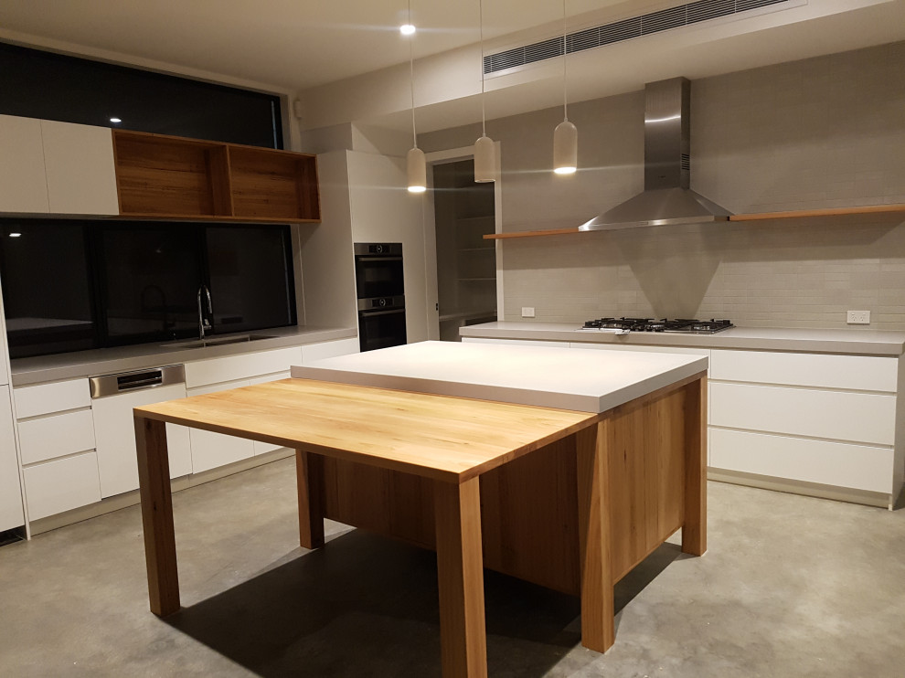 Large contemporary l-shaped eat-in kitchen in Geelong with a double-bowl sink, flat-panel cabinets, white cabinets, concrete benchtops, grey splashback, ceramic splashback, panelled appliances, concrete floors, with island and grey benchtop.