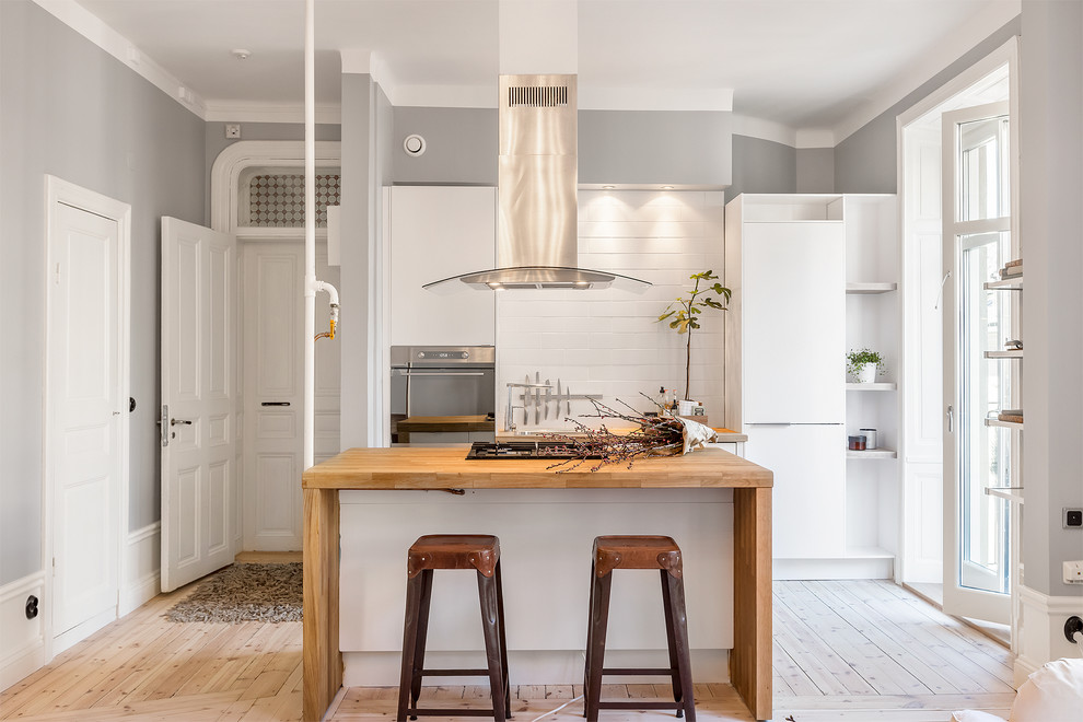 Inspiration for a mid-sized scandinavian galley open plan kitchen in Stockholm with flat-panel cabinets, white cabinets, wood benchtops, white splashback, subway tile splashback, stainless steel appliances, light hardwood floors and with island.