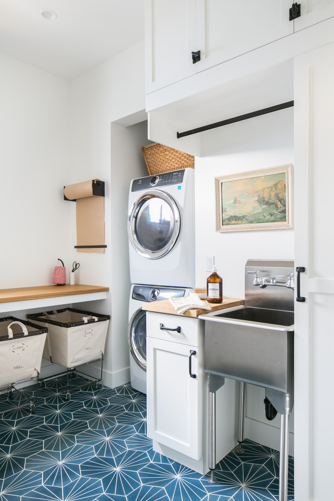 Beach style laundry room in Orange County with an utility sink, orange cabinets, wood benchtops, white walls, a stacked washer and dryer, blue floor and brown benchtop.
