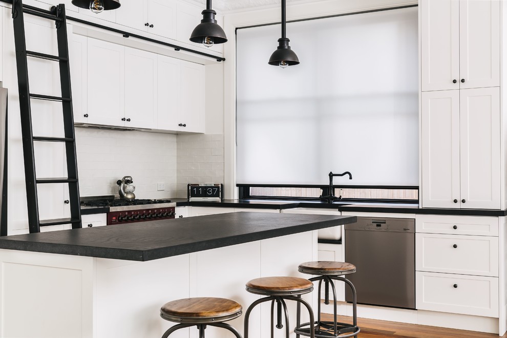 Photo of a large industrial l-shaped kitchen in Sydney with a farmhouse sink, shaker cabinets, white cabinets, white splashback, stainless steel appliances, medium hardwood floors, with island, brown floor and black benchtop.