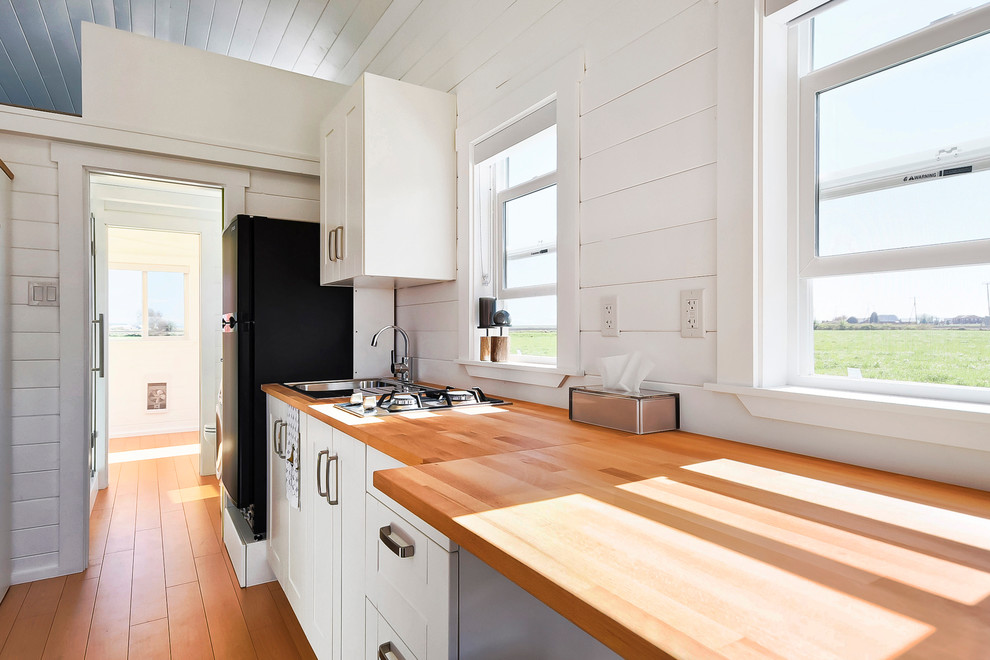 Small contemporary single-wall eat-in kitchen in Vancouver with a drop-in sink, flat-panel cabinets, white cabinets, wood benchtops, black appliances, light hardwood floors and with island.