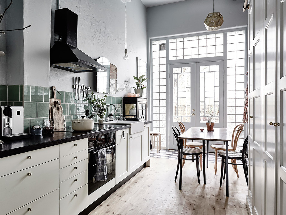 Inspiration for a mid-sized scandinavian single-wall eat-in kitchen in Gothenburg with a farmhouse sink, flat-panel cabinets, white cabinets, black appliances, light hardwood floors, wood benchtops, green splashback, porcelain splashback and no island.