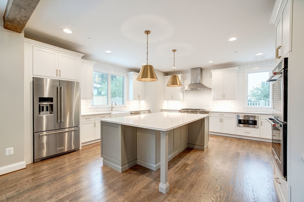 This is an example of a country l-shaped open plan kitchen in DC Metro with a farmhouse sink, shaker cabinets, white cabinets, solid surface benchtops, white splashback, subway tile splashback, stainless steel appliances, medium hardwood floors, with island, brown floor and white benchtop.