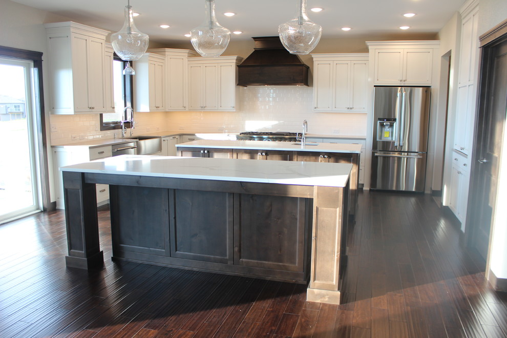 Large transitional l-shaped dark wood floor enclosed kitchen photo with a farmhouse sink, shaker cabinets, white cabinets, quartz countertops, white backsplash, subway tile backsplash, stainless steel appliances and an island