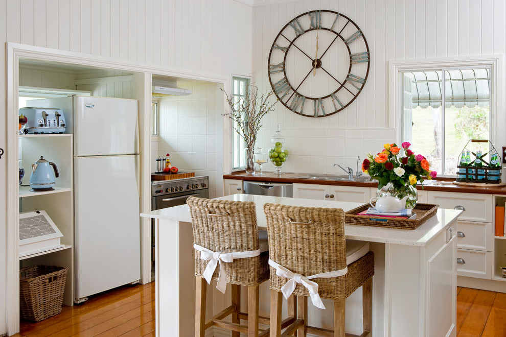 Beach style u-shaped kitchen in Brisbane with a drop-in sink, recessed-panel cabinets, white cabinets, white splashback, stainless steel appliances, medium hardwood floors, with island, brown floor and white benchtop.