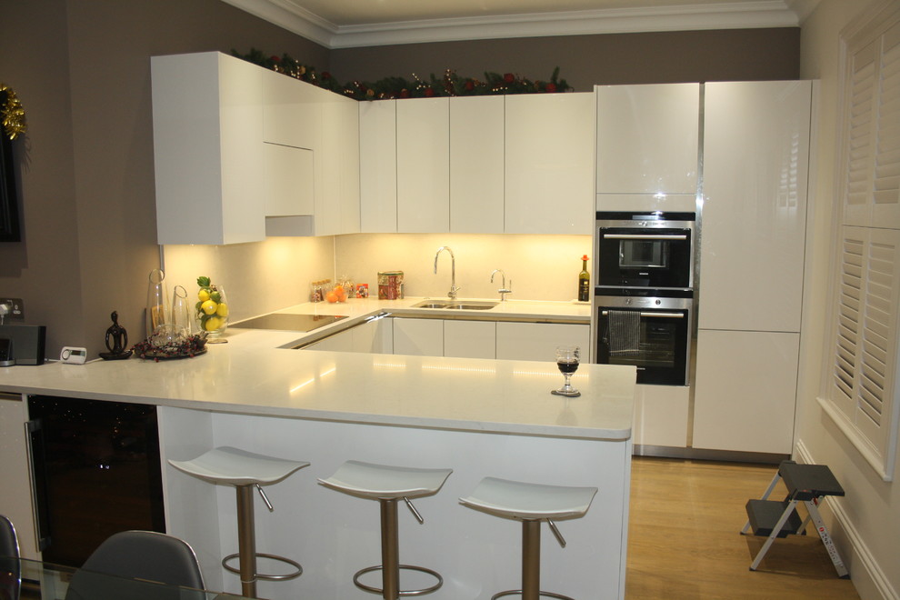 This is an example of a large modern u-shaped open plan kitchen in London with an undermount sink, flat-panel cabinets, white cabinets, quartzite benchtops, white splashback, black appliances, medium hardwood floors and a peninsula.