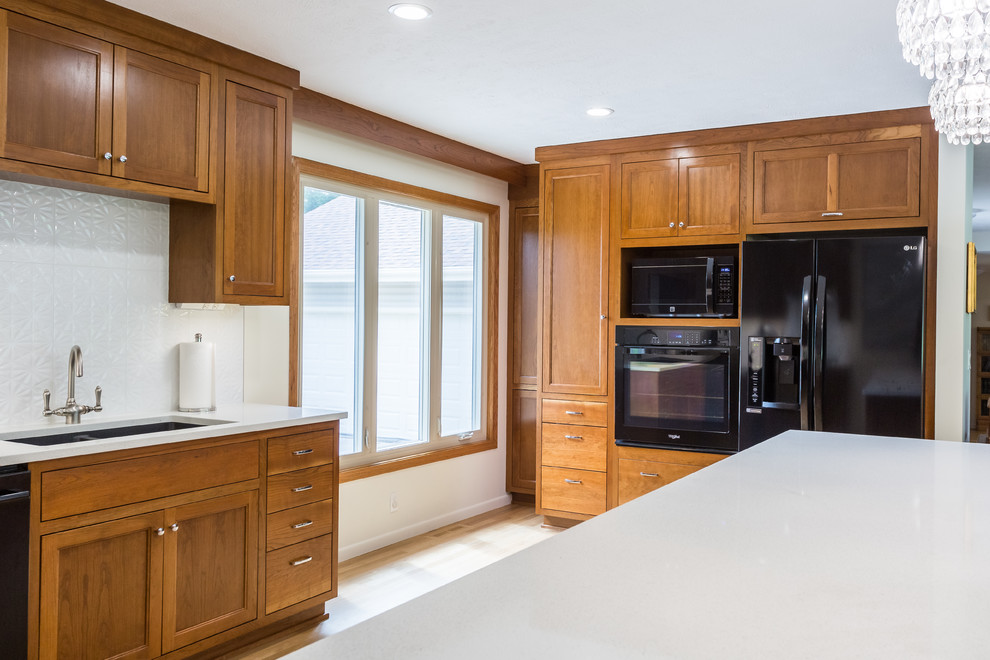 This is an example of a mid-sized midcentury l-shaped eat-in kitchen in Omaha with an undermount sink, flat-panel cabinets, medium wood cabinets, quartz benchtops, white splashback, porcelain splashback, black appliances, light hardwood floors, with island, white floor and white benchtop.
