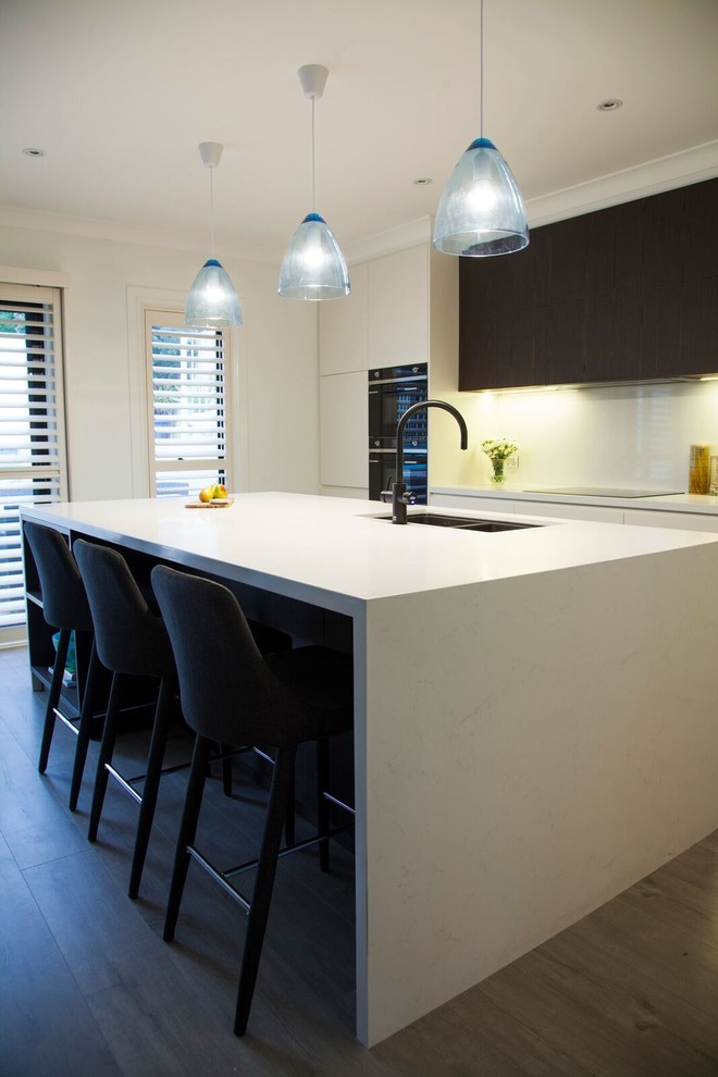 This is an example of a mid-sized contemporary galley open plan kitchen in Sydney with a double-bowl sink, flat-panel cabinets, white cabinets, quartz benchtops, white splashback, stone slab splashback, black appliances, light hardwood floors and with island.